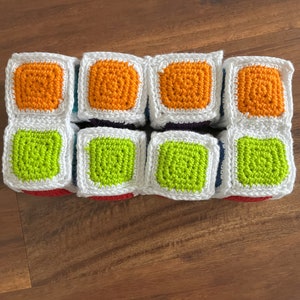 May include: Nine crocheted squares in a 3x3 grid. The squares are white with a colored square in the center. The colors are orange, green, and purple.