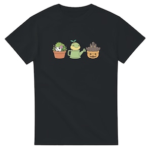 May include: Black t-shirt featuring a whimsical illustration of three potted plants with cute animal faces. The plants include a hedgehog, a watering can with a frog, and a pumpkin with a cat. The design is in a cartoon style.