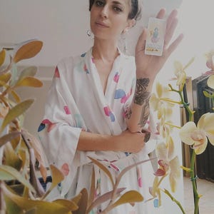 Puede incluir: Una bata blanca con un patr&oacute;n abstracto colorido. Se sostiene una carta del tarot, con una ilustraci&oacute;n detallada. El fondo incluye plantas con flores amarillas. La bata est&aacute; abierta, revelando un vistazo de la persona que la usa.