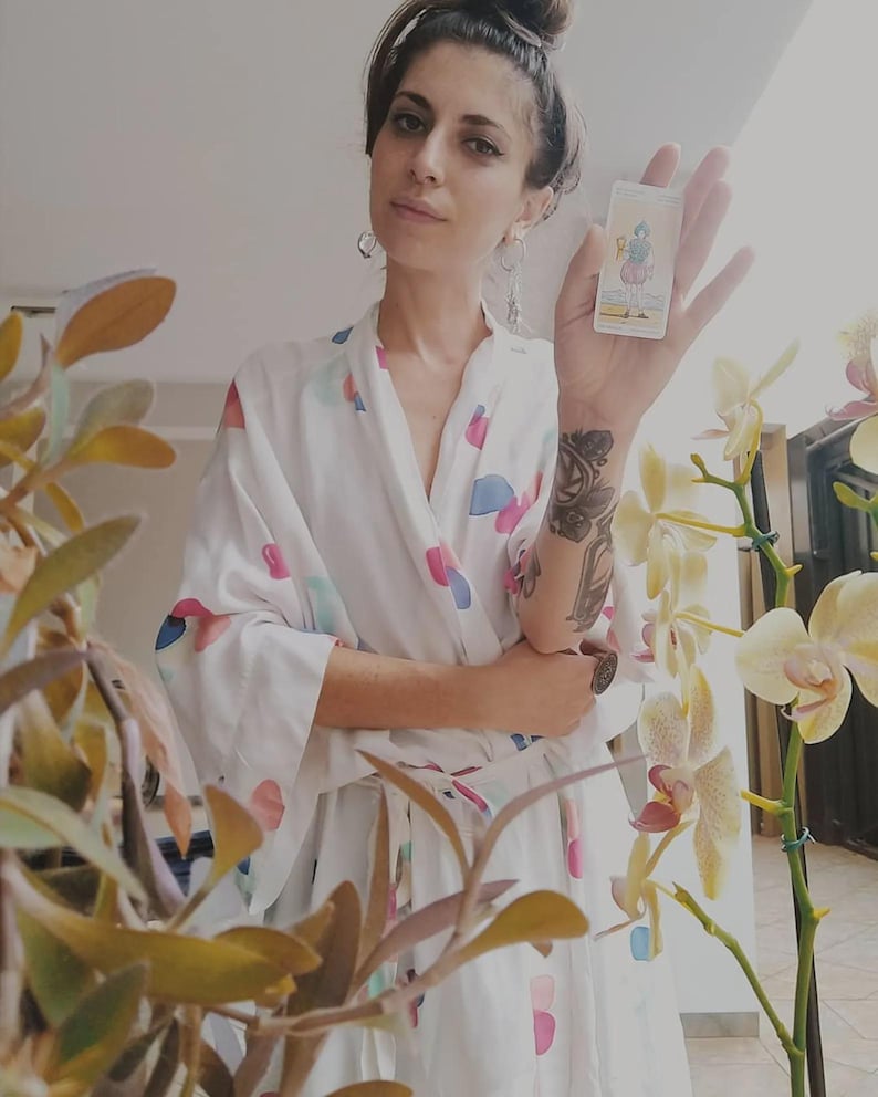Puede incluir: Una bata blanca con un patr&oacute;n abstracto colorido. Se sostiene una carta del tarot, con una ilustraci&oacute;n detallada. El fondo incluye plantas con flores amarillas. La bata est&aacute; abierta, revelando un vistazo de la persona que la usa.