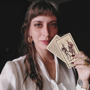Puede incluir: Una persona sostiene dos cartas del tarot. Las cartas tienen un fondo beige con ilustraciones de figuras con ropa hist&oacute;rica. La persona lleva una blusa blanca y pendientes dorados. El fondo es oscuro.
