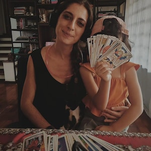 May include: A woman and child are holding tarot cards. The woman is wearing a black top and headphones. The child is wearing an orange top and a pink cap. A deck of tarot cards is spread out on a patterned surface.