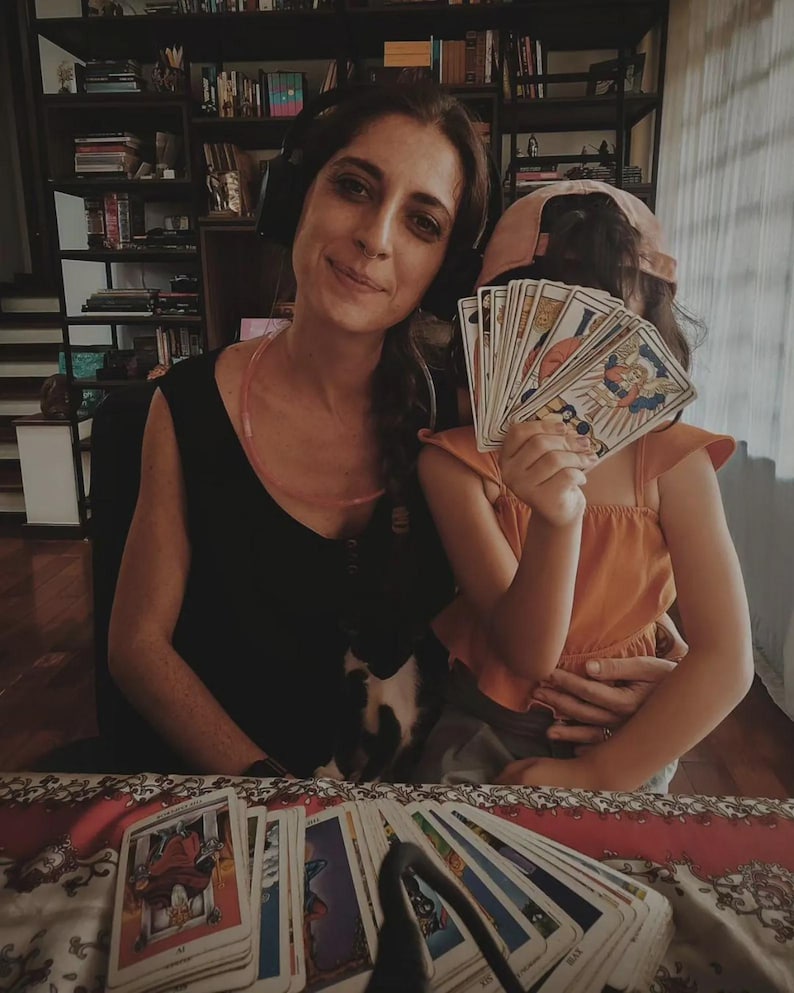 May include: A woman and child are holding tarot cards. The woman is wearing a black top and headphones. The child is wearing an orange top and a pink cap. A deck of tarot cards is spread out on a patterned surface.