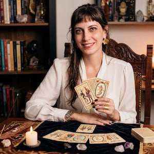 Puede incluir: Una persona sonriendo mientras sostiene cartas del tarot. Las cartas son de color beige con ilustraciones y texto. Una vela encendida, incienso y otras cartas del tarot est&aacute;n sobre la mesa. La mesa est&aacute; cubierta con una tela azul oscuro.