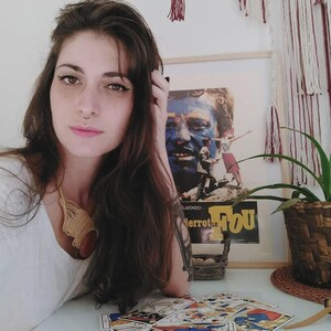 Puede incluir: Una mujer con cabello casta&ntilde;o largo y un piercing en la nariz est&aacute; sentada en una mesa con cartas del tarot. Un p&oacute;ster de pel&iacute;cula enmarcado es visible en el fondo. Se usa un collar tejido.