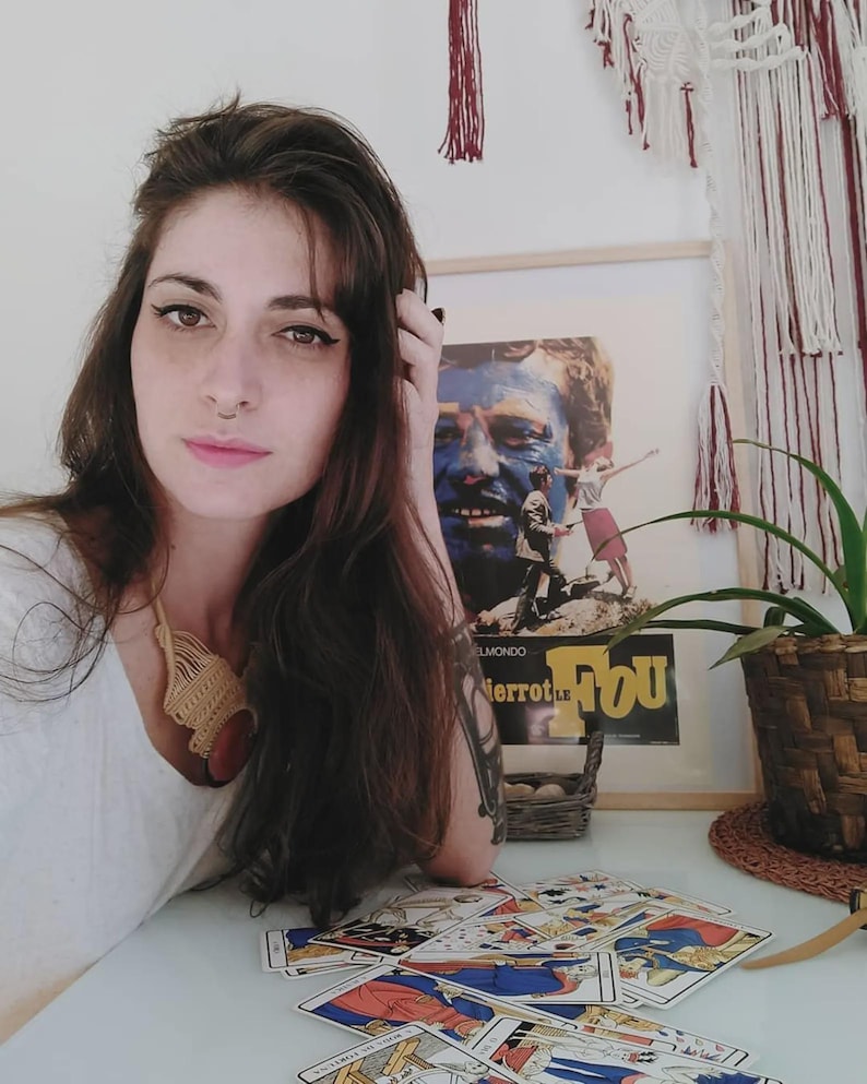Puede incluir: Una mujer con cabello casta&ntilde;o largo y un piercing en la nariz est&aacute; sentada en una mesa con cartas del tarot. Un p&oacute;ster de pel&iacute;cula enmarcado es visible en el fondo. Se usa un collar tejido.