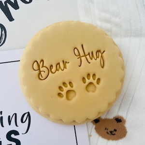 May include: A circular, pale brown biscuit with a decorative edge. The words "Bear Hug" and two paw prints are stamped on the biscuit. The biscuit rests on a white surface with a small bear graphic and the word "smile" in the background.