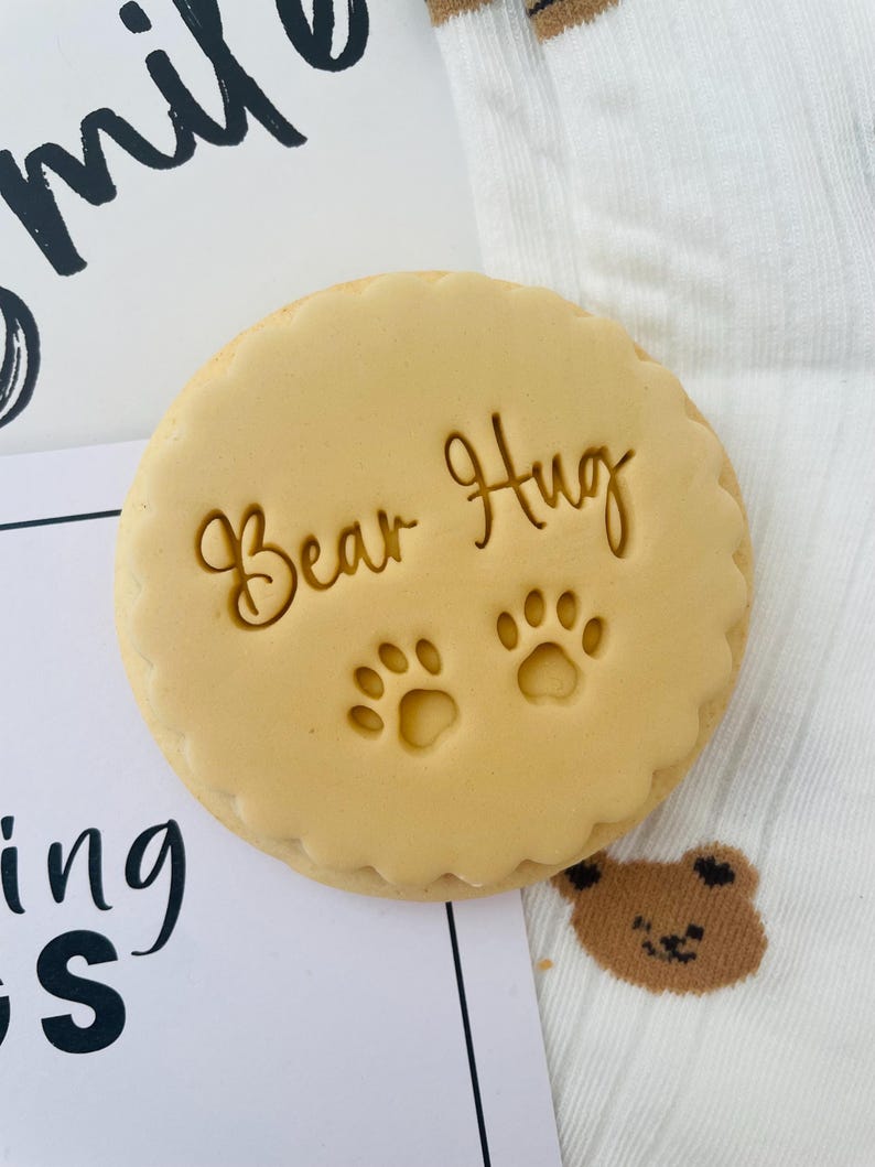 May include: A circular, pale brown biscuit with a decorative edge. The words "Bear Hug" and two paw prints are stamped on the biscuit. The biscuit rests on a white surface with a small bear graphic and the word "smile" in the background.