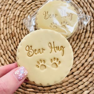 Pu&ograve; includere: Due biscotti rotondi di colore marrone chiaro con il testo "Bear Hug" e due impronte di zampe in glassa dorata.