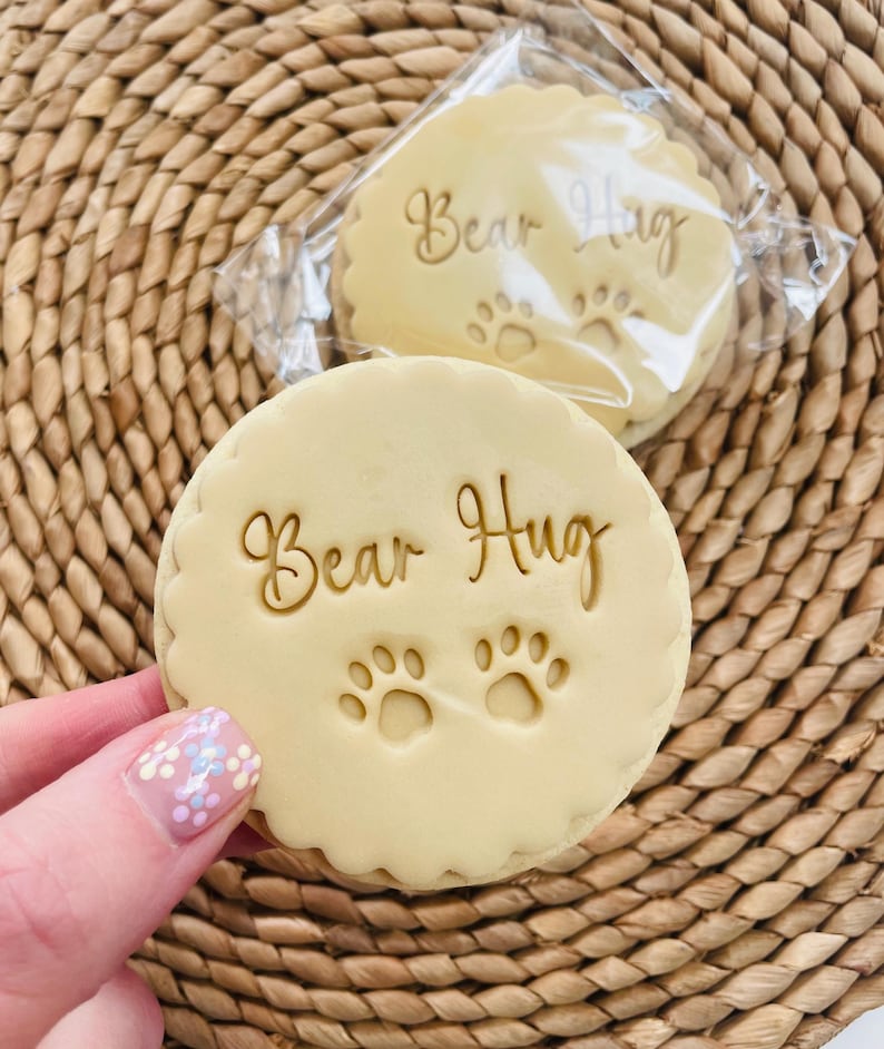 Pu&ograve; includere: Due biscotti rotondi di colore marrone chiaro con il testo "Bear Hug" e due impronte di zampe in glassa dorata.