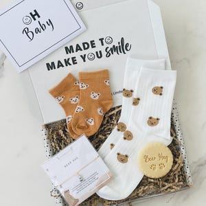 Pu&ograve; includere: Una scatola regalo piena di trucioli di carta marrone e un biglietto bianco con il testo "Made to make you smile". La scatola contiene un paio di calzini bianchi con facce di orsi marroni, un paio di calzini marroni con facce di orsi bianchi, un piccolo biglietto bianco con il testo "Bear Hug" e un braccialetto d'argento con un ciondolo a forma di orso.