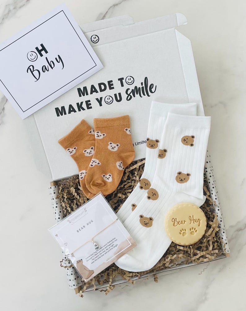 Pu&ograve; includere: Una scatola regalo piena di trucioli di carta marrone e un biglietto bianco con il testo "Made to make you smile". La scatola contiene un paio di calzini bianchi con facce di orsi marroni, un paio di calzini marroni con facce di orsi bianchi, un piccolo biglietto bianco con il testo "Bear Hug" e un braccialetto d'argento con un ciondolo a forma di orso.