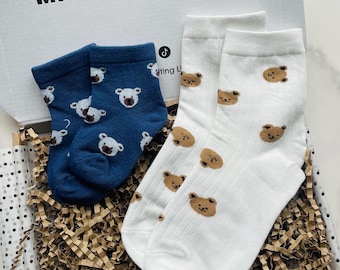 May include: A gift box containing two pairs of socks. One pair is navy blue with white bear faces, and the other is white with brown bear faces. The box has the text "MADE TO MAKE YOU smile".