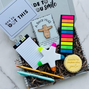May include: A gift box filled with motivational items for students. The box includes a notepad, a pencil, a pen, a star-shaped highlighter, a wooden bear hug cutout, a sticky note pad, and a cookie that says "Made to make you smile".