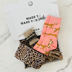May include: A gift box filled with a pink and brown leopard print pouch, a pair of pink socks with a cheetah print, a brown burlap pouch with gold and black beaded bracelets, and a brown hair clip. The box is white with the text "MADE TO MAKE YOU smile".