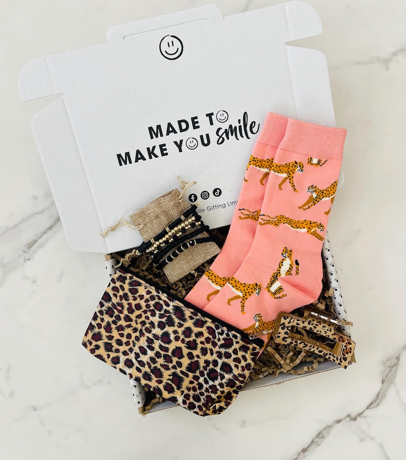 May include: A gift box filled with a pink and brown leopard print pouch, a pair of pink socks with a cheetah print, a brown burlap pouch with gold and black beaded bracelets, and a brown hair clip. The box is white with the text "MADE TO MAKE YOU smile".