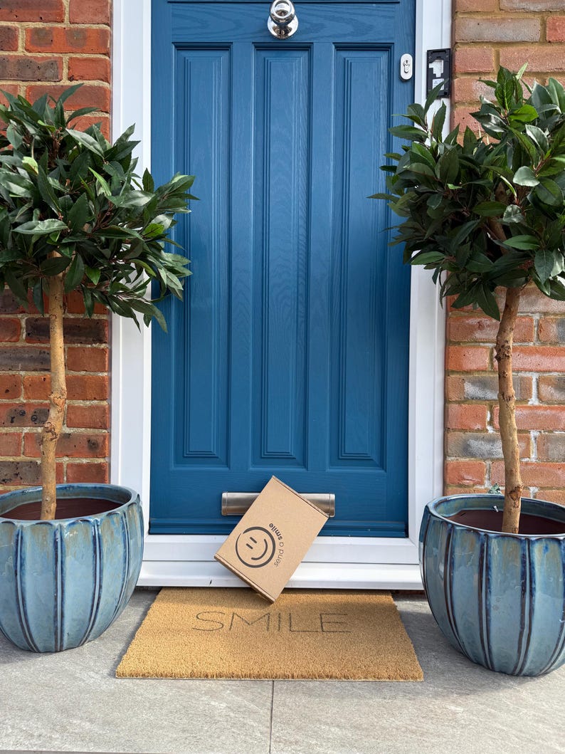May include: A teal blue front door with a silver door knocker and mail slot. A package with a smiley face logo rests on a doormat that reads "SMILE". Two potted trees flank the door, set against a brick wall.