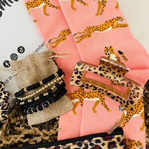 May include: A pink and brown leopard print gift box filled with a pair of pink socks with a leopard print, a brown leopard print hair clip, a small brown burlap bag with black and gold beaded bracelets, and a leopard print makeup bag.