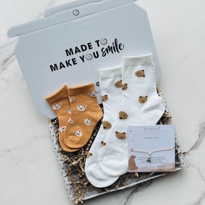 Pu&ograve; includere: Una scatola regalo bianca con la scritta "Made to make you smile" stampata sulla parte superiore. La scatola contiene due paia di calzini con stampa di orso, un paio &egrave; bianco con orsi marroni e l'altro paio &egrave; arancione con orsi marroni. La scatola contiene anche un piccolo biglietto con la scritta "Bear Hug" e un braccialetto con un ciondolo a forma di orso.