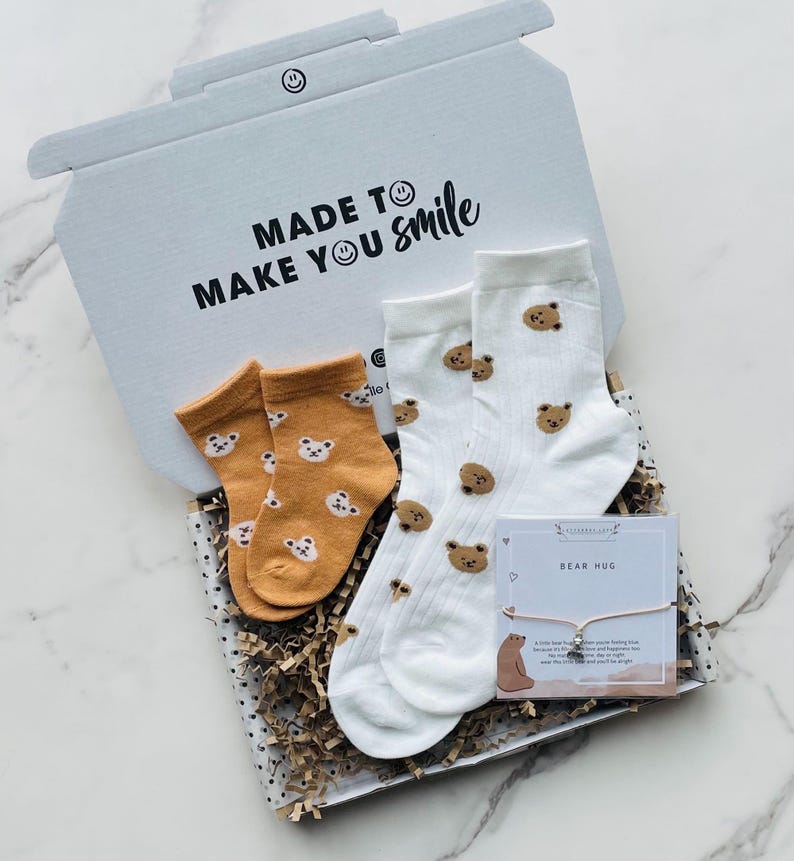 Pu&ograve; includere: Una scatola regalo bianca con la scritta "Made to make you smile" stampata sulla parte superiore. La scatola contiene due paia di calzini con stampa di orso, un paio &egrave; bianco con orsi marroni e l'altro paio &egrave; arancione con orsi marroni. La scatola contiene anche un piccolo biglietto con la scritta "Bear Hug" e un braccialetto con un ciondolo a forma di orso.