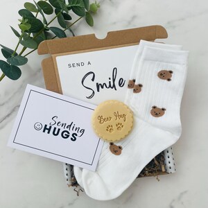 May include: A gift box filled with a white card that says "Sending Hugs" with a smiley face, a white pair of socks with brown bear designs, and a round, light brown cookie with the words "Bear Hug" and paw prints.