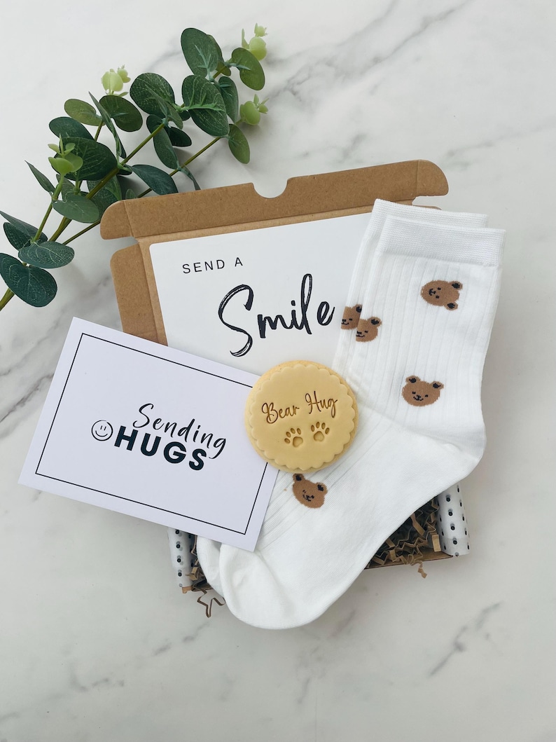 May include: A gift box filled with a white card that says "Sending Hugs" with a smiley face, a white pair of socks with brown bear designs, and a round, light brown cookie with the words "Bear Hug" and paw prints.