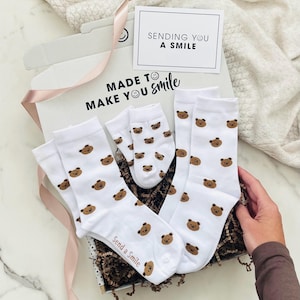 May include: A white gift box containing three pairs of white socks with a brown bear face pattern. The box lid features the text "MADE TO MAKE YOU smile" and a card reading "SENDING YOU A SMILE". One sock pair has "Send a Smile" printed on the cuff.