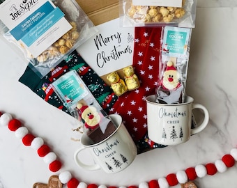 May include: A Christmas gift box filled with treats, including two bags of gourmet popcorn, a mug, hot chocolate stirrer, socks, and chocolate candies. The box is decorated with a "Merry Christmas" sign and a red and white garland.