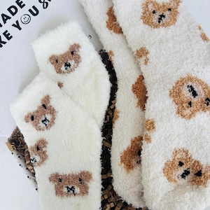May include: A pair of fluffy white socks and another pair of white socks with brown teddy bear faces are displayed in a gift box. The text "MADE TO MAKE YOU SMILE" is visible on the box. These cosy socks are ideal for gifting.