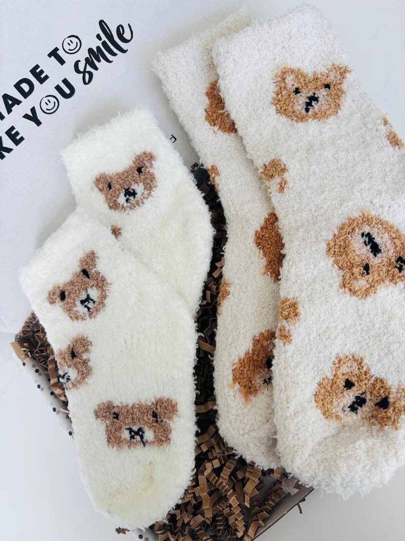 May include: A pair of fluffy white socks and another pair of white socks with brown teddy bear faces are displayed in a gift box. The text "MADE TO MAKE YOU SMILE" is visible on the box. These cosy socks are ideal for gifting.