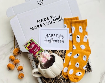 May include: A Halloween gift box containing a white pumpkin mug with hot chocolate and marshmallows, orange ghost-patterned socks, and a card reading "Happy Halloween". The box also includes a bar of chocolate and the words "Made to Make You Smile".
