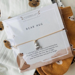 Pu&ograve; includere: Un biglietto bianco con il testo "LETTERBOX LOVE" e "BEAR HUG" su di esso. Il biglietto &egrave; accanto a un paio di calzini bianchi con stampe di orsi marroni e un paio di calzini marroni con stampe di orsi marroni. Un braccialetto con un piccolo ciondolo a forma di orso &egrave; sul biglietto. Il braccialetto &egrave; beige e ha un messaggio sul biglietto che dice "Un piccolo abbraccio di orso per quando ti senti gi&ugrave;, perch&eacute; &egrave; pieno di amore e felicit&agrave;. Non importa l'ora, il giorno o la notte, indossa questo piccolo orso e starai bene."