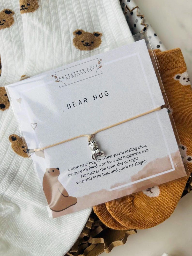 Pu&ograve; includere: Un biglietto bianco con il testo "LETTERBOX LOVE" e "BEAR HUG" su di esso. Il biglietto &egrave; accanto a un paio di calzini bianchi con stampe di orsi marroni e un paio di calzini marroni con stampe di orsi marroni. Un braccialetto con un piccolo ciondolo a forma di orso &egrave; sul biglietto. Il braccialetto &egrave; beige e ha un messaggio sul biglietto che dice "Un piccolo abbraccio di orso per quando ti senti gi&ugrave;, perch&eacute; &egrave; pieno di amore e felicit&agrave;. Non importa l'ora, il giorno o la notte, indossa questo piccolo orso e starai bene."