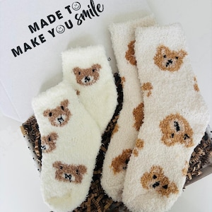 May include: A set of fluffy white socks with brown teddy bear faces, presented in an open gift box. The box lid has the words "MADE TO MAKE YOU smile" printed on it.