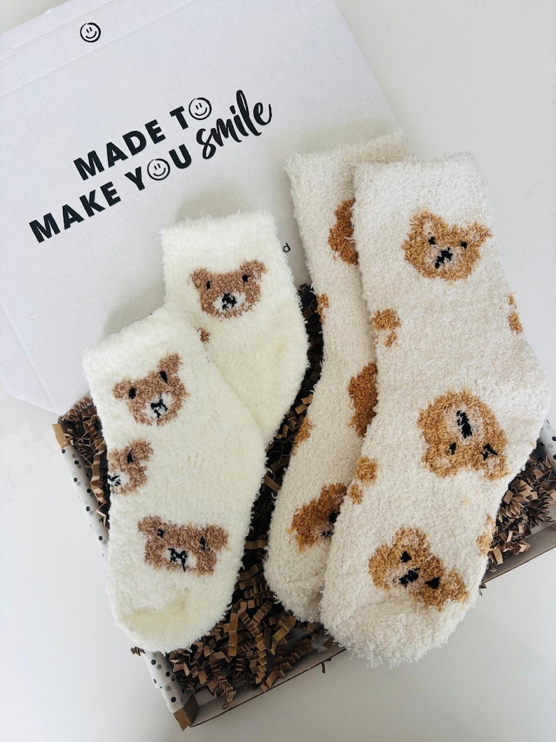 May include: A set of fluffy white socks with brown teddy bear faces, presented in an open gift box. The box lid has the words "MADE TO MAKE YOU smile" printed on it.