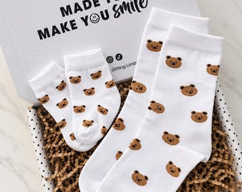 May include: A pair of white socks and a pair of smaller white socks, both featuring a repeating brown bear face pattern. The socks are presented in a white cardboard gift box with the text MADE TO MAKE YOU smile printed on the inside lid.