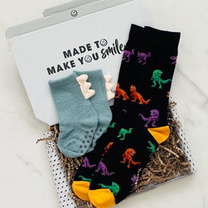 May include: A gift box with two pairs of socks. One pair is black with colourful dinosaur graphics, and the other is light blue with a dinosaur design. The box has the text "MADE TO MAKE YOU smile".
