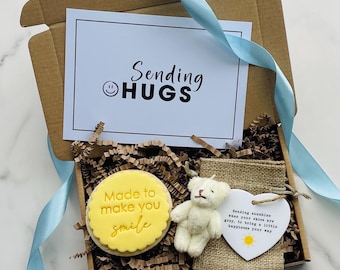 May include: A brown gift box tied with a light blue ribbon, containing a card with "Sending HUGS", a yellow cookie with "Made to make you smile", a small teddy bear, and a heart-shaped tag with a message.