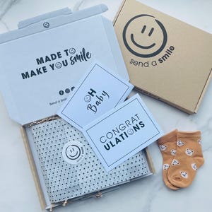 Pu&ograve; includere: Una scatola bianca con il testo "MADE TO MAKE YOU Smile" e un logo con una faccina sorridente. All'interno della scatola c'&egrave; carta velina bianca con pois neri. Ci sono due biglietti, uno che dice "OH Baby" con una faccina sorridente e l'altro che dice "CONGRATULATIONS". Ci sono anche due paia di calzini per neonati arancioni con una stampa di orso.