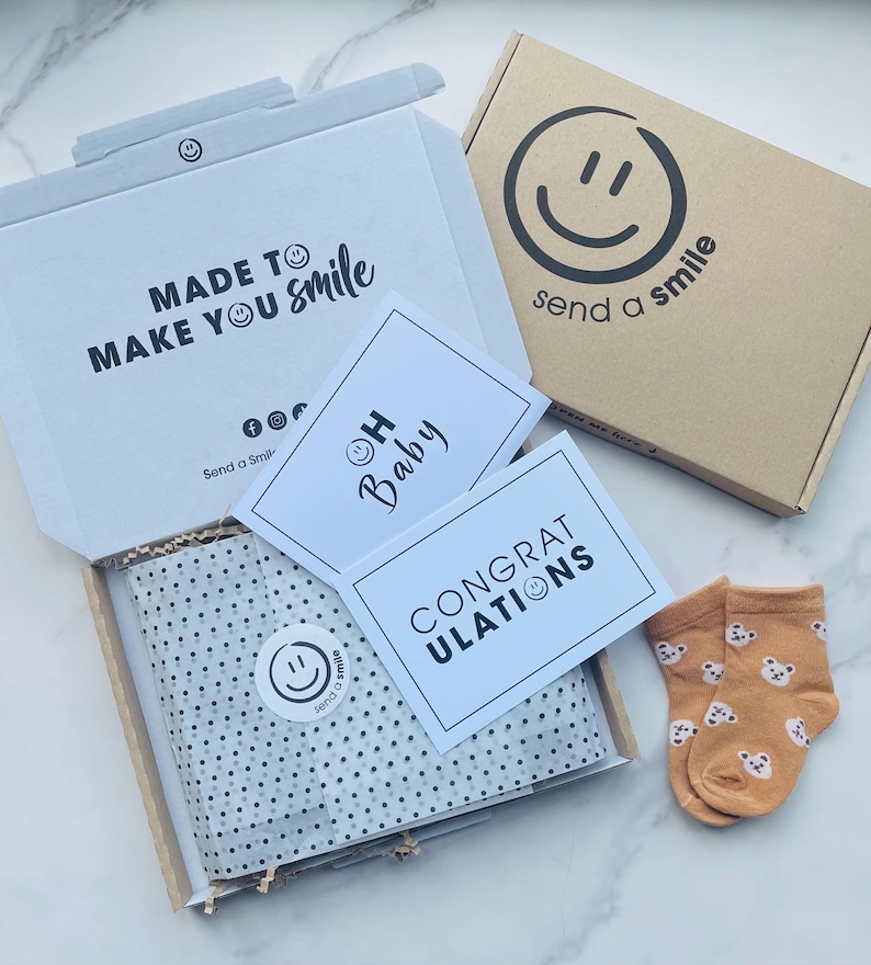 Pu&ograve; includere: Una scatola bianca con il testo "MADE TO MAKE YOU Smile" e un logo con una faccina sorridente. All'interno della scatola c'&egrave; carta velina bianca con pois neri. Ci sono due biglietti, uno che dice "OH Baby" con una faccina sorridente e l'altro che dice "CONGRATULATIONS". Ci sono anche due paia di calzini per neonati arancioni con una stampa di orso.