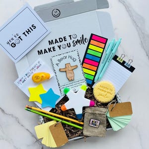 May include: A gift box containing stationery items, including a card with the text "you've GOT THIS", pens, sticky notes, pencils, and a small bag. The box also has the text "MADE TO MAKE YOU smile".