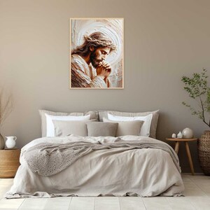 May include: A painting of Jesus Christ with a crown of thorns, praying with his hands clasped together. The painting is in a light brown frame and is hanging on a beige wall.