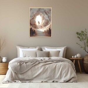 Puede incluir: Una pintura que representa la apertura de una cueva con una gran piedra rodada, revelando una luz brillante y una figura caminando hacia ella. La pintura est&aacute; enmarcada y colgada en una pared sobre una cama con un edred&oacute;n de lino beige.