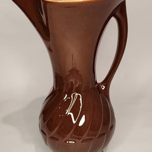 May include: A brown ceramic pitcher with a wide mouth and a curved handle. The pitcher has a textured, ribbed design.