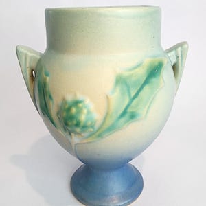 May include: A blue and green glazed ceramic vase with two handles and a floral design. The vase has a wide mouth and a round body with a pedestal base.