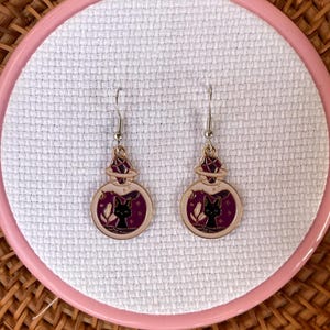 May include: Dangle earrings with a round pendant showing a black cat silhouette in a purple and white potion bottle design. The earrings have silver hooks. The background is white fabric.