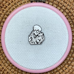 May include: A black and white enamel pin featuring a skeleton holding a small cat, centred on a piece of white Aida fabric stretched within a round, light pink plastic embroidery hoop. The design shows the skeleton in profile embracing the cat.