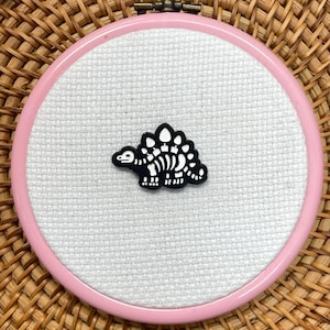 May include: A pink embroidery hoop holds white fabric with a black and white dinosaur skeleton design. The hoop is set against a woven basket background, creating a contrast of textures and colours.