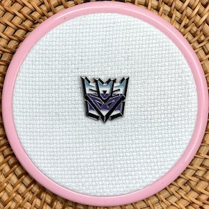 May include: A Transformers Decepticon logo pin, featuring a purple, black, and blue design. The pin is centered on white cross-stitch fabric, set within a pink embroidery hoop. The background is a woven wicker basket.
