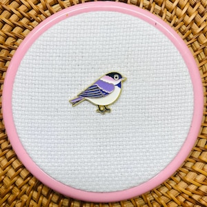 Peut inclure: Une broche en émail métallique en forme de petit oiseau avec une poitrine blanche, une aile violette à rayures bleu foncé et une tache rose clair sur la tête. La broche est centrée sur un tissu blanc circulaire dans un cercle en plastique rose.